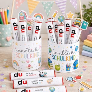 May include: Two white cylindrical containers decorated with colorful school-themed illustrations and the text "endlich SCHULKIND". Each container holds several white chocolate bars labeled "du" with various school-themed toppers and text.