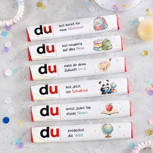 May include: Six rectangular chocolate bars with white wrappers, each featuring the word 'du' in black and red, German text, and colorful illustrations including a backpack, books, paint palette, panda, apple, and globe, arranged vertically.