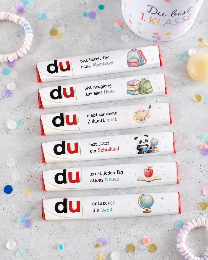 May include: Six rectangular chocolate bars with white wrappers, each featuring the word 'du' in black and red, German text, and colorful illustrations including a backpack, books, paint palette, panda, apple, and globe, arranged vertically.