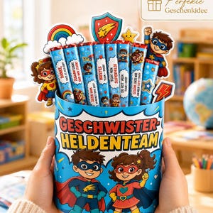 Könnte beinhalten: Ein zylindrischer Behälter im Superhelden-Design mit der Aufschrift "GESCHWISTER-HELDENTEAM", gefüllt mit mehreren Schokoriegeln. Jeder Riegel zeigt Superhelden-Illustrationen und deutschen Text. Dekorative Superhelden-Aufsteller sind angebracht.