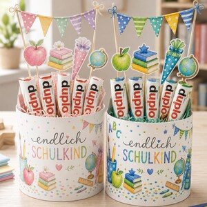 May include: Two cylindrical gift boxes labeled "endlich SCHULKIND" filled with Duplo chocolate bars. Each box features school-themed illustrations like apples, books, and globes, topped with decorative paper pennant banners and cutouts of school supplies.