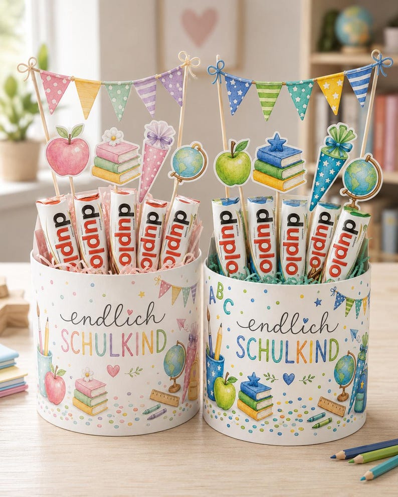 May include: Two cylindrical gift boxes labeled "endlich SCHULKIND" filled with Duplo chocolate bars. Each box features school-themed illustrations like apples, books, and globes, topped with decorative paper pennant banners and cutouts of school supplies.