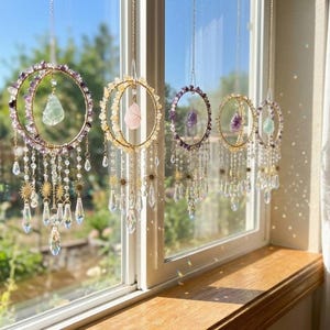 Puede incluir: Una colección de atrapasoles de cristal colgantes. Cada atrapasol presenta un anillo de metal dorado o plateado adornado con cristales y piedras preciosas, con cuentas de cristal colgantes y detalles de estrellas. Los atrapasoles se exhiben frente a una ventana.