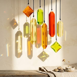 Puede incluir: Adornos de vidrio facetado de colores, suspendidos de cuerdas negras. Los colores incluyen naranja, amarillo, verde y rojo. Los adornos proyectan sombras sobre una pared blanca, creando un juego de luces. Formas: diamantes y rectángulos.