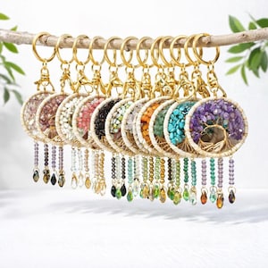 May include: A collection of keychains featuring a tree of life design. Each keychain has a gold-colored metal ring and a circular pendant filled with colorful gemstones. The pendants are adorned with beaded accents and hanging crystal charms.