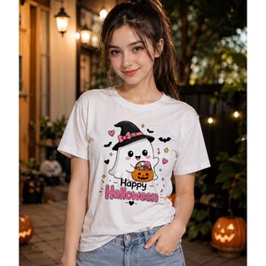 May include: A white short-sleeve t-shirt featuring a graphic of a cartoon ghost wearing a black witch hat and holding a jack-o'-lantern bucket filled with candy. The text "Happy Halloween" is printed in pink below the ghost with small bats.
