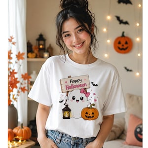 May include: A white short-sleeved t-shirt featuring a graphic of a smiling ghost with a pink bow, holding a wooden sign that reads Happy Halloween. The design includes a lit lantern, a jack-o'-lantern, small pink hearts, and black bats.