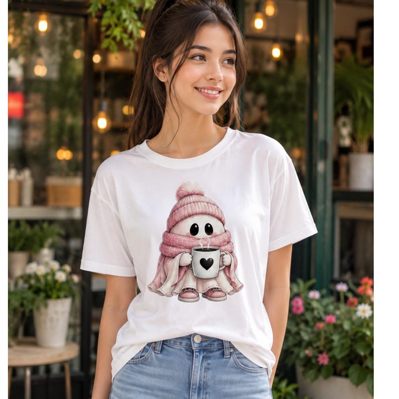May include: A white short-sleeve t-shirt featuring a graphic of a small white ghost character wearing a pink knit beanie and a matching pink scarf. The ghost is holding a white mug with a black heart design and wearing pink sneakers.