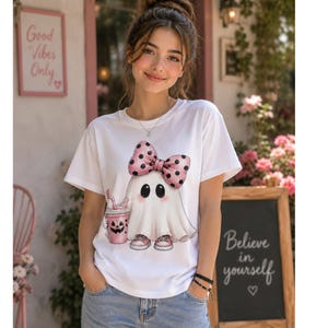 May include: A white short-sleeved t-shirt featuring a graphic of a ghost wearing a pink polka-dot bow and pink sneakers, holding a pink jack-o'-lantern cup with a straw. The ghost has large black eyes and rosy cheeks on a white fabric base.