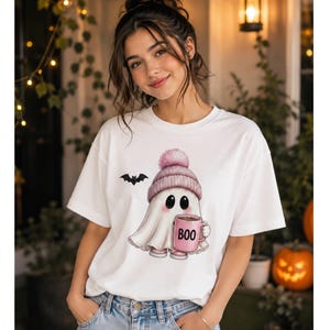 May include: A white t-shirt featuring a graphic of a ghost wearing a pink knit hat with a pom-pom, holding a pink mug with the text BOO printed on it. A small black bat is flying to the left of the ghost. The design is centered on the chest.