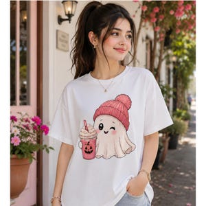 May include: A white short-sleeved t-shirt featuring a graphic illustration of a cute ghost wearing a pink knit beanie with a pom-pom. The ghost is holding a pink iced coffee cup decorated with a black jack-o'-lantern face and a straw.
