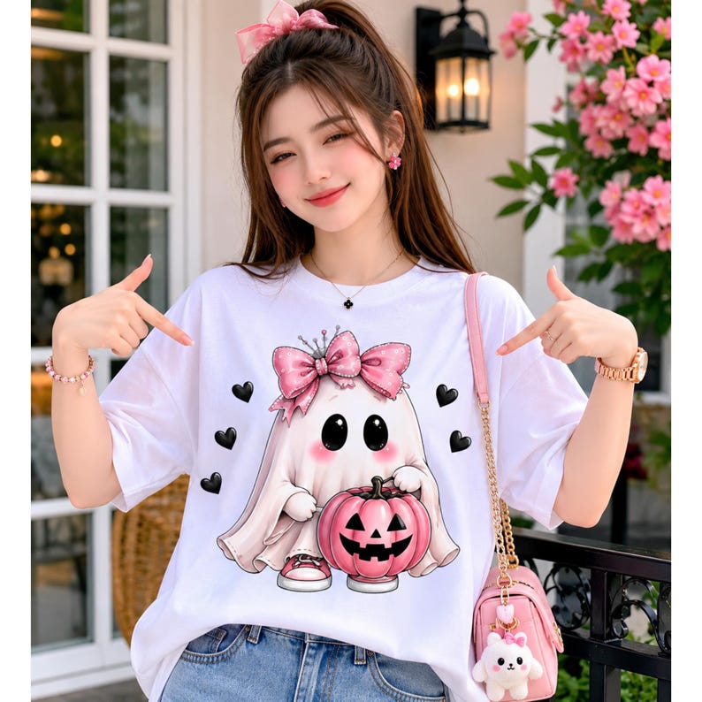 May include: A white short-sleeved t-shirt featuring a graphic of a cartoon ghost wearing a large pink polka-dot bow. The ghost holds a pink jack-o'-lantern pumpkin and is surrounded by small black hearts. The design is centered on the chest.