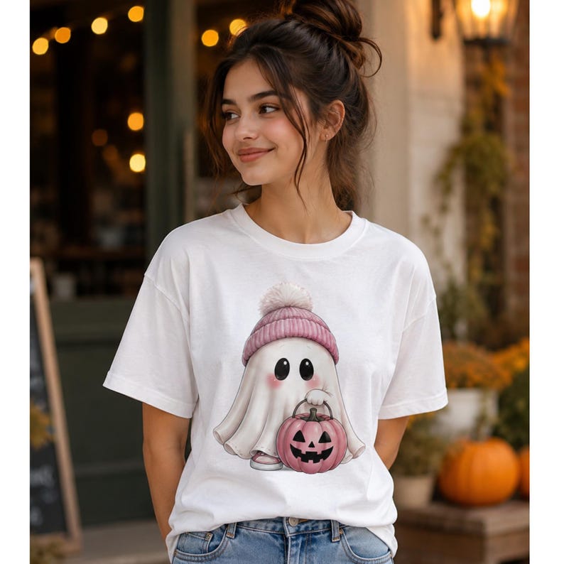 May include: A white short-sleeved t-shirt featuring a graphic of a white ghost wearing a pink knit beanie with a pom-pom. The ghost has rosy cheeks and is holding a pink jack-o'-lantern pumpkin bucket with a black carved face design.