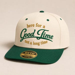 May include: A cream-colored baseball cap with a dark green brim and embroidered text that reads here for a Good Time not a long time. The cap features a circular gold and black sticker on the brim with the text OTTO WORLD FAMOUS PREMIUM OSFM.