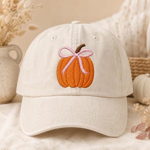 May include: A cream-colored baseball cap featuring an embroidered design of an orange pumpkin with a brown stem and a light pink ribbon tied in a bow around the top. The cap has a structured crown, a curved brim, and stitched eyelet details.