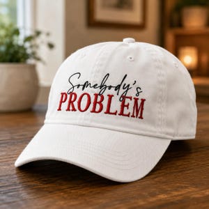 Puede incluir: Una gorra de béisbol blanca con texto bordado en la parte frontal. La palabra Somebody's está cosida en una fuente cursiva negra, mientras que la palabra PROBLEM está cosida debajo en una fuente roja, negrita y con serifa en mayúsculas.