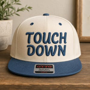 Op de afbeelding: Een crèmekleurige snapback-pet met de tekst TOUCH DOWN in donkerblauw geborduurd op de voorzijde. De pet heeft een platte donkerblauwe klep met een rechthoekige sticker met de tekst OTTO PREMIUM FABRIC FLAT BILL SNAPBACK OSFM in zwart en rood.