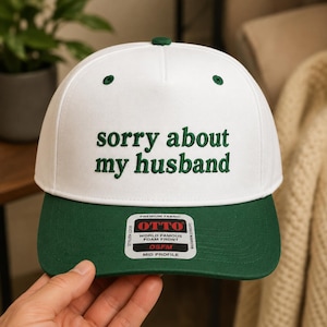 May include: A white baseball cap with a dark green brim and dark green embroidered text on the front that reads sorry about my husband. The cap features a rectangular sticker on the brim with the text OTTO, PREMIUM FABRIC, and OSFM.