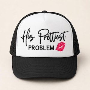 Könnte beinhalten: Eine weiß-schwarze Trucker-Cap mit schwarzer Stickerei in Schreibschrift mit der Aufschrift His Prettiest und in Blockschrift mit der Aufschrift PROBLEM, ergänzt durch einen kleinen, pinkfarbenen Kussmund als Stickerei.