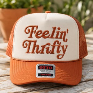 May include: A rust-orange and cream trucker hat featuring the embroidered text Feelin' Thrifty in a cursive font on the front foam panel. The hat includes a mesh back, a curved brim, and a rectangular sticker on the brim reading OTTO.