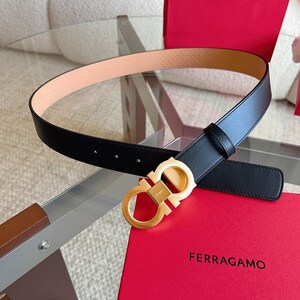 May include: Black leather belt with a gold-tone buckle. The belt has a light brown interior and is displayed on a glass surface next to a red box with the brand name "FERRAGAMO".