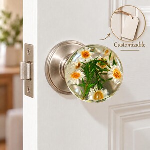 May include: A round door knob with a clear resin finish containing embedded white and yellow daisy-like flowers and green foliage. The knob features a brushed nickel base and latch plate. A small circular inset shows a blank tag labeled Customizable.