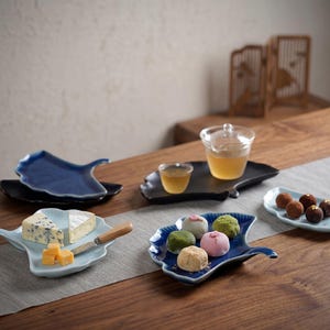 Puede incluir: Una colección de platos de cerámica con forma de hoja en azul, negro y azul claro. Los platos contienen queso, mochi y trufas. También hay un vaso de té y un cuchillo pequeño.