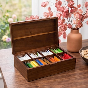 May include: A dark wooden tea box with an open lid, showing compartments filled with colourful tea bags. The box is on a wooden surface. A vase with pink flowers is in the background. The tea bags are in various colours.