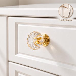 Peut inclure: Un bouton de tiroir en verre transparent à facettes avec une base en métal doré, monté sur un tiroir d'armoire blanc. Le bouton présente un design circulaire nervuré qui capte la lumière, et un encart circulaire affiche une étiquette blanche avec le texte Customizable.