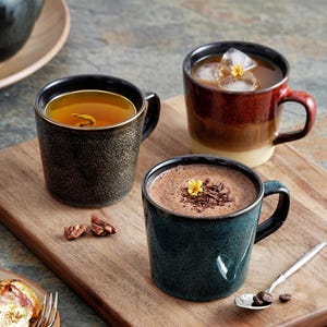 Peut inclure: Trois tasses en céramique contenant des boissons sont posées sur une planche en bois. L'une est vert foncé avec du thé, une autre est rouge et marron avec du café glacé, et la troisième est bleu sarcelle avec du chocolat chaud.