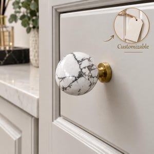May include: A spherical cabinet knob featuring a white surface with dark grey veining, mounted on a circular brushed brass base. The knob is attached to a light grey drawer front. A circular inset shows a blank tag with the text Customizable.