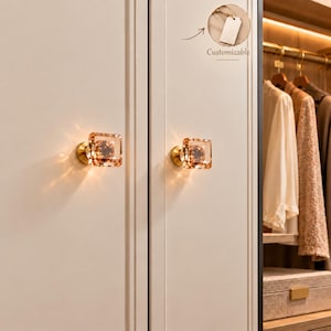 May include: Two square, amber-colored crystal cabinet knobs with gold-toned metal bases mounted on white cabinet doors. A circular inset at the top right shows a blank white tag with the text Customizable, indicating the product is customizable.