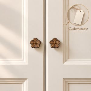 Pode incluir: Dois puxadores de armário em forma de flor, em tom de bronze, com cinco pétalas e um detalhe circular central, montados num par de portas de armário brancas com painéis. Uma etiqueta retangular em branco apresenta o texto Customizable.