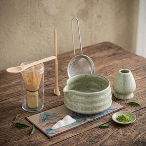 May include: A matcha tea set on a wooden surface. The set includes a green ceramic bowl, a whisk, a scoop, a sifter, a small dish with green powder, and a bamboo spoon. A small ceramic stand and a decorative mat are also included.