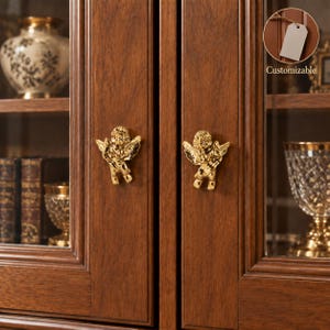 May include: Two gold-colored metal cabinet knobs shaped like cherubs with wings, mounted on a dark brown wooden cabinet door. The knobs feature a shiny, reflective finish. A small circular inset in the top right corner shows a blank tag and the text Customizable.