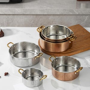 Peut inclure: Un ensemble de quatre casseroles rondes en acier inoxydable avec un motif de grille texturé sur l'extérieur. Chaque casserole possède deux petites poignées en métal doré sur les côtés opposés. Les casseroles sont de tailles variées.