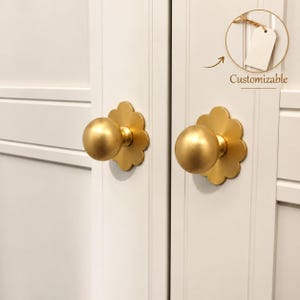 May include: Two matte gold spherical cabinet knobs mounted on white paneled doors. Each knob features a scalloped, flower-shaped backplate. A circular graphic in the upper right corner shows a blank white tag with the text Customizable.