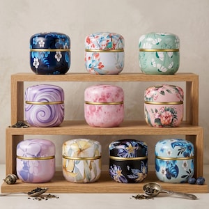 May include: A wooden shelf displays nine small, round metal tins with floral designs. The tins feature various colors, including blue, pink, purple, and green, with gold-colored bands. Some tins have swirling patterns or watercolor-style flowers.
