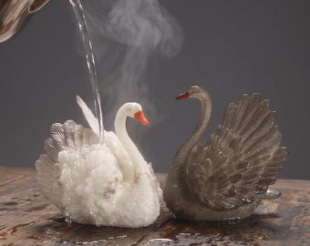 May include: Two swan-shaped figurines, one white and one dark gray, with detailed feather textures. A metal pitcher pours water over the white swan, creating steam. The white swan has an orange beak, while the gray swan has a dark beak.