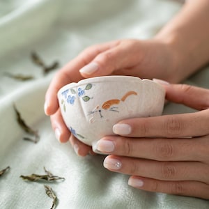 May include: A small, off-white ceramic bowl with a scalloped rim. The bowl is decorated with blue floral accents and a playful orange and white cat design. The bowl is held in two hands.