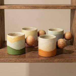May include: Four ceramic mugs with cylindrical bodies and spherical wooden handles. The mugs feature varied color-blocked designs in shades of yellow, green, pink, blue, and orange, arranged in horizontal bands or layered patterns on the exterior.