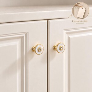 Peut inclure: Deux boutons de placard ronds avec un bord en métal doré perlé, une incrustation en nacre blanche et un cristal central transparent. Les boutons sont fixés sur des portes de placard blanc cassé. Une icône d'étiquette indique Customizable.