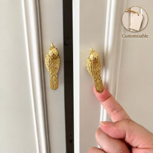 Puede incluir: Dos pomos de armario de metal dorado con forma de pájaro y texturas detalladas de alas y plumas. Los pomos están montados en puertas de armario blancas. Un pequeño recuadro circular en la esquina superior derecha muestra una etiqueta con el texto Customizable.
