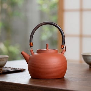 May include: A terracotta-colored teapot with a dark wooden handle and a small spout. The teapot has a rounded body with horizontal ridges and a small lid with a knob. The handle is attached with copper-colored metal. A traditional tea set.