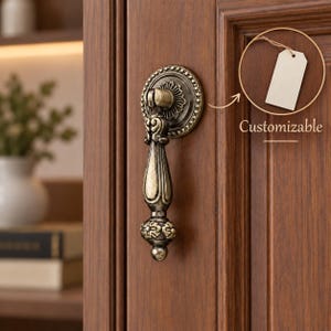May include: A bronze-toned metal drop pull handle with an ornate, textured circular backplate mounted on a wooden cabinet door. The handle features a decorative, elongated design with floral-like detailing and the text Customizable.