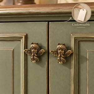 May include: Two brass-toned metal cabinet knobs shaped like winged figures with hands held together, mounted on sage green cabinet doors. A circular inset shows a blank white tag with the text Customizable.