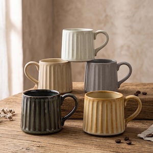 May include: A set of five ceramic mugs with a vertical ribbed texture and speckled finish. The mugs are arranged in a stack and group, featuring colors of off-white, beige, grey, dark green, and golden brown, each with a curved side handle.