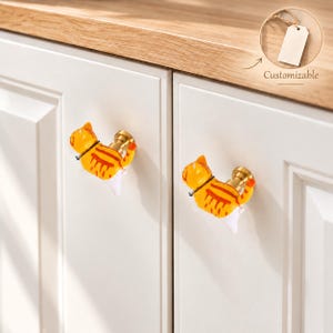 May include: Two cabinet knobs shaped like orange tabby cats with white bellies and brass-colored hardware. The cats are depicted in a climbing position, attached to white cabinet doors. A small circular inset shows a blank tag labeled Customizable.