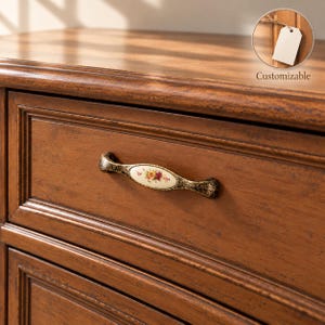 Puede incluir: Un cajón de madera marrón con un tirador metálico decorativo con un centro de cerámica ovalado. El tirador tiene un acabado oscuro y desgastado con un patrón floral en la pieza de cerámica color crema. Un recuadro circular muestra una etiqueta vacía con el texto Customizable.