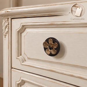 May include: A cream-coloured wooden dresser drawer featuring a decorative drawer pull shaped like a five-petaled flower with a dark circular base and a metallic gold finish. An inset circle shows a blank white tag with the text Customizable.